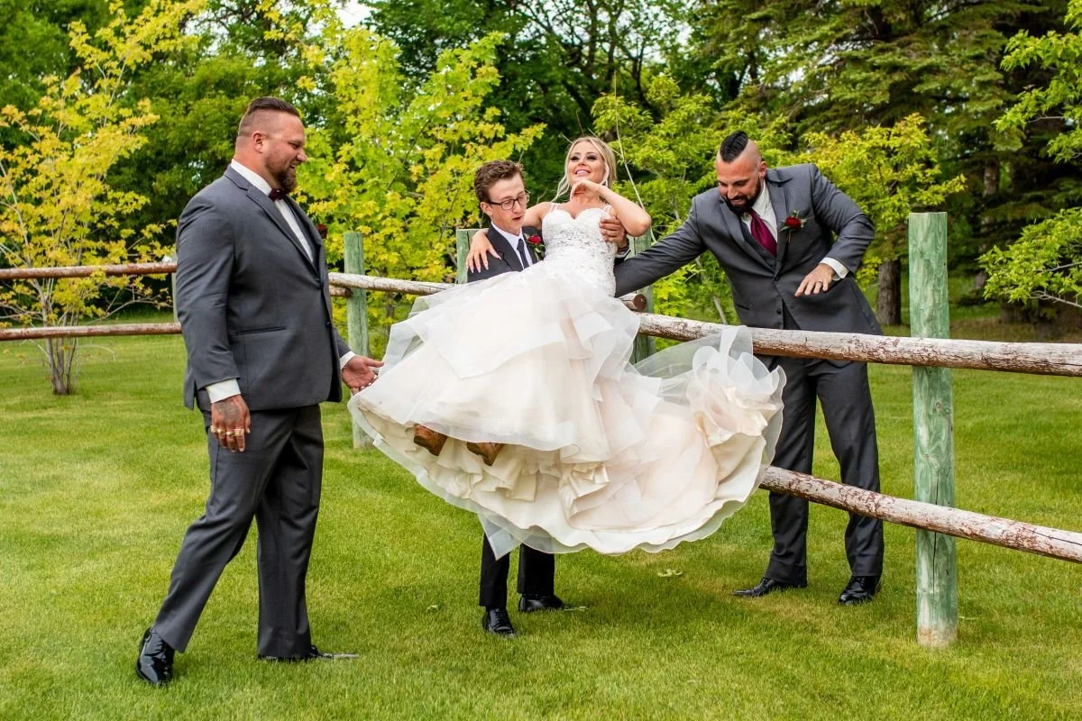 A funny and candid wedding party photo of a groom and groomsmen in grey suits lifting up a laughing bride in her ruffled mermaid gown next to a rustic wooden fence at the Hitch 'n Post venue near Winnipeg.