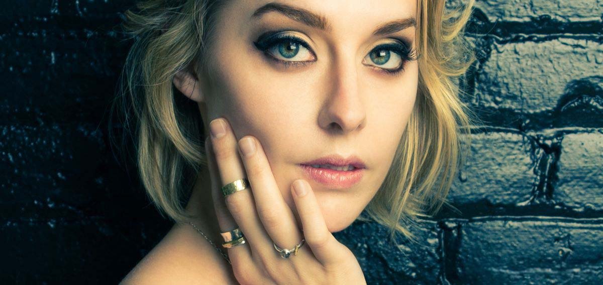 A close-up editorial beauty portrait of a blonde woman with dramatic smokey eyeliner and gold rings, resting her hand on her cheek against a dark painted brick wall in a photography studio in Winnipeg, Manitoba.