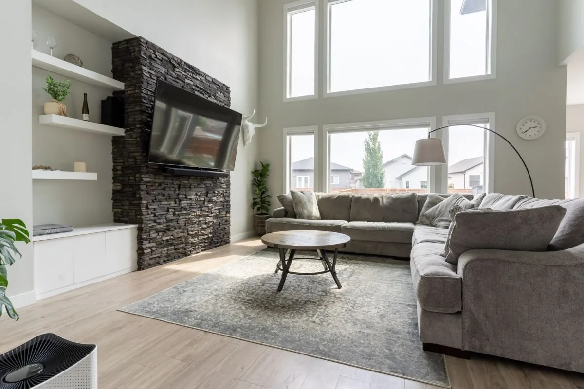 A professional real estate listing photograph of a luxury living room featuring a stacked stone fireplace and double-height windows, shot for a RE/MAX agent by a Winnipeg real estate photographer.