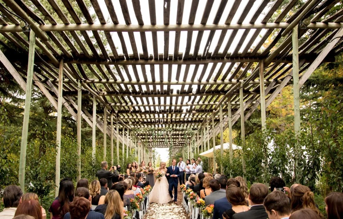 A wide-angle shot of a bride in a beige tulle maxi dress and a groom in a rich navy blue suit walking down an outdoor wedding aisle lined with guests under a massive rustic wooden pergola at The Saskatoon Farm venue during an Alberta destination wedd