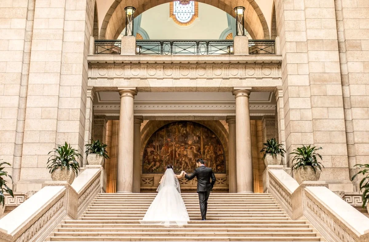 A wide-angle wedding portrait of a bride and groom holding hands while walking up the grand staircase under the historic arches of the Manitoba Legislative Building.