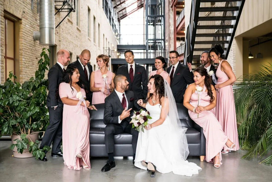 A bride wearing a white dress and veil sits beside a groom in a dark grey suit with a burgundy tie and pocket square, surrounded by 10 wedding party members at the Exchange District Campus of RRC Polytech in Winnipeg, Manitoba.