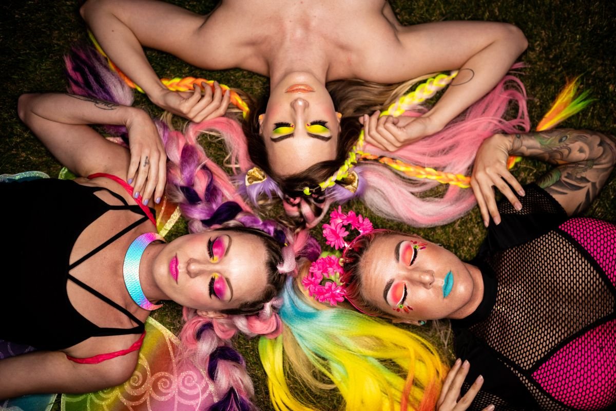 Three women in vibrant makeup and colourful rave outfits with rainbow hair lie in the grass with their heads touching.