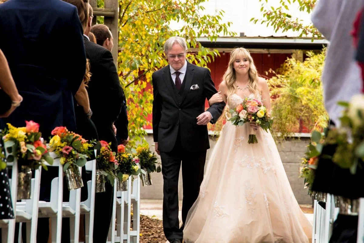 A bride in a beige tulle maxi dress is walked down the outdoor ceremony aisle by her father in a black suit at The Saskatoon Farm venue during a destination wedding in Alberta, captured by a Winnipeg wedding photographer.