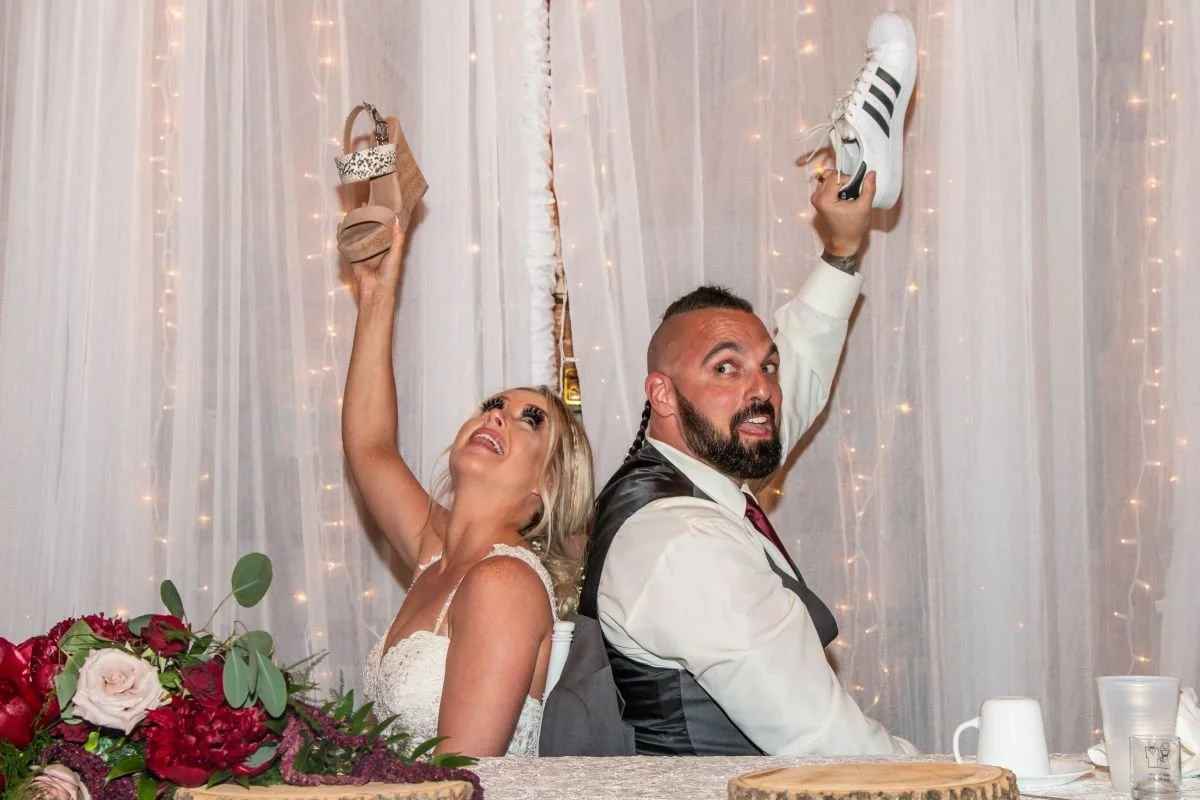A candid close-up of a bride and groom sitting back-to-back playing the wedding shoe game, with the bride raising her wedge shoe and the groom holding up a white sneaker against a backdrop of fairy string lights.