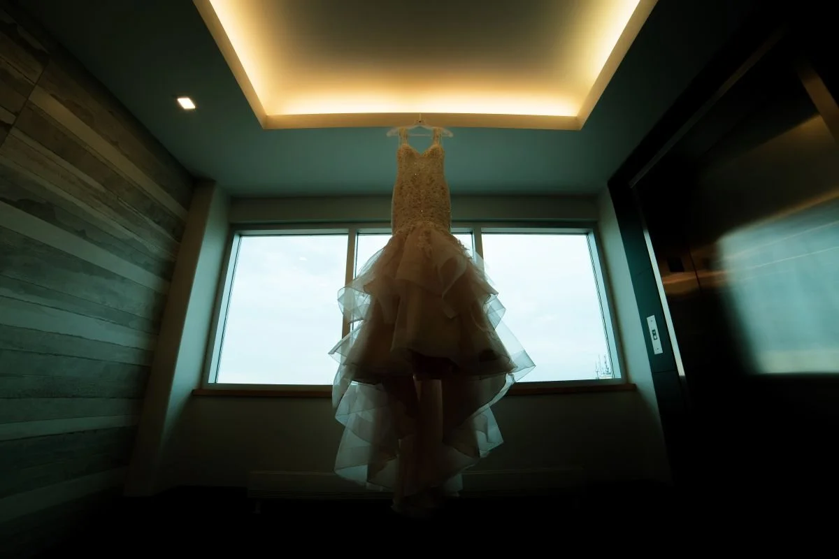 A wedding gown with a lace bodice and a dramatic layered ruffle mermaid skirt hanging in a window frame under warm recessed tray lighting.