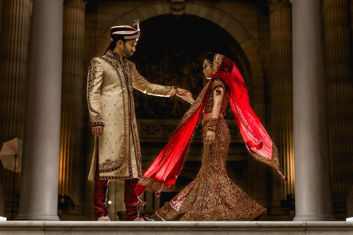A dramatic evening portrait of a groom in a gold sherwani holding hands with his bride in an embroidered red and gold lehenga under the stone arches of the Manitoba Legislative Building.