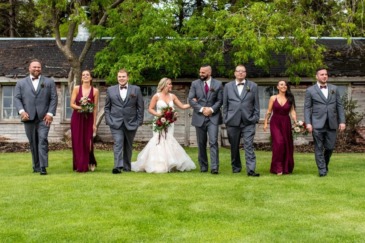 A wide-angle wedding party photo of a bride and groom walking across a grass lawn with bridesmaids in burgundy dresses and groomsmen in grey suits in front of a rustic wooden barn.