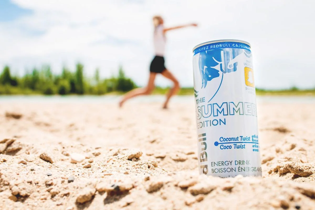A sharp product shot of a Red Bull Summer Edition Coconut Twist energy drink can standing on a sandy beach, with a blurred model running in the background; click to view the commercial and product photography gallery