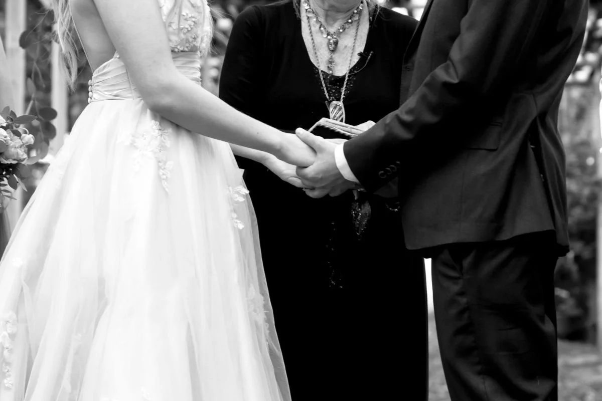 A  black and white photo of a bride holding hands with a groom in front of an officiant during a destination wedding session at The Saskatoon Farm in Foothills County, Alberta by a Winnipeg wedding photographer.