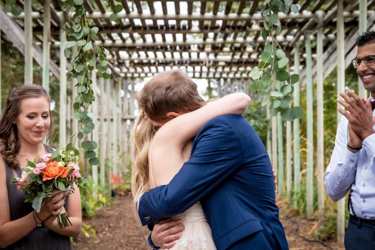 A close-up candid of a bride and groom embracing under hanging eucalyptus greens at The Saskatoon Farm venue, captured during an Alberta destination wedding by a Winnipeg wedding photographer.
