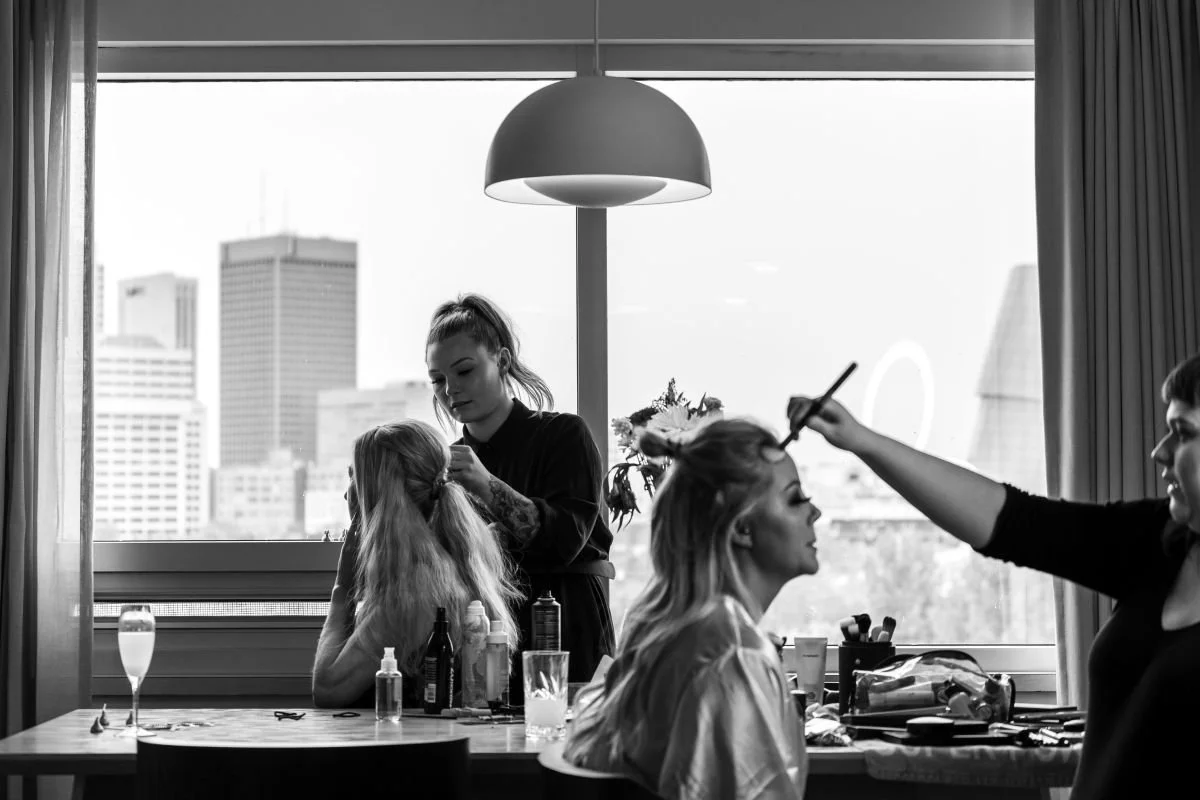 A black and white candid photo of a bride getting her makeup and hair styled by professionals in front of a window overlooking the Winnipeg city skyline.
