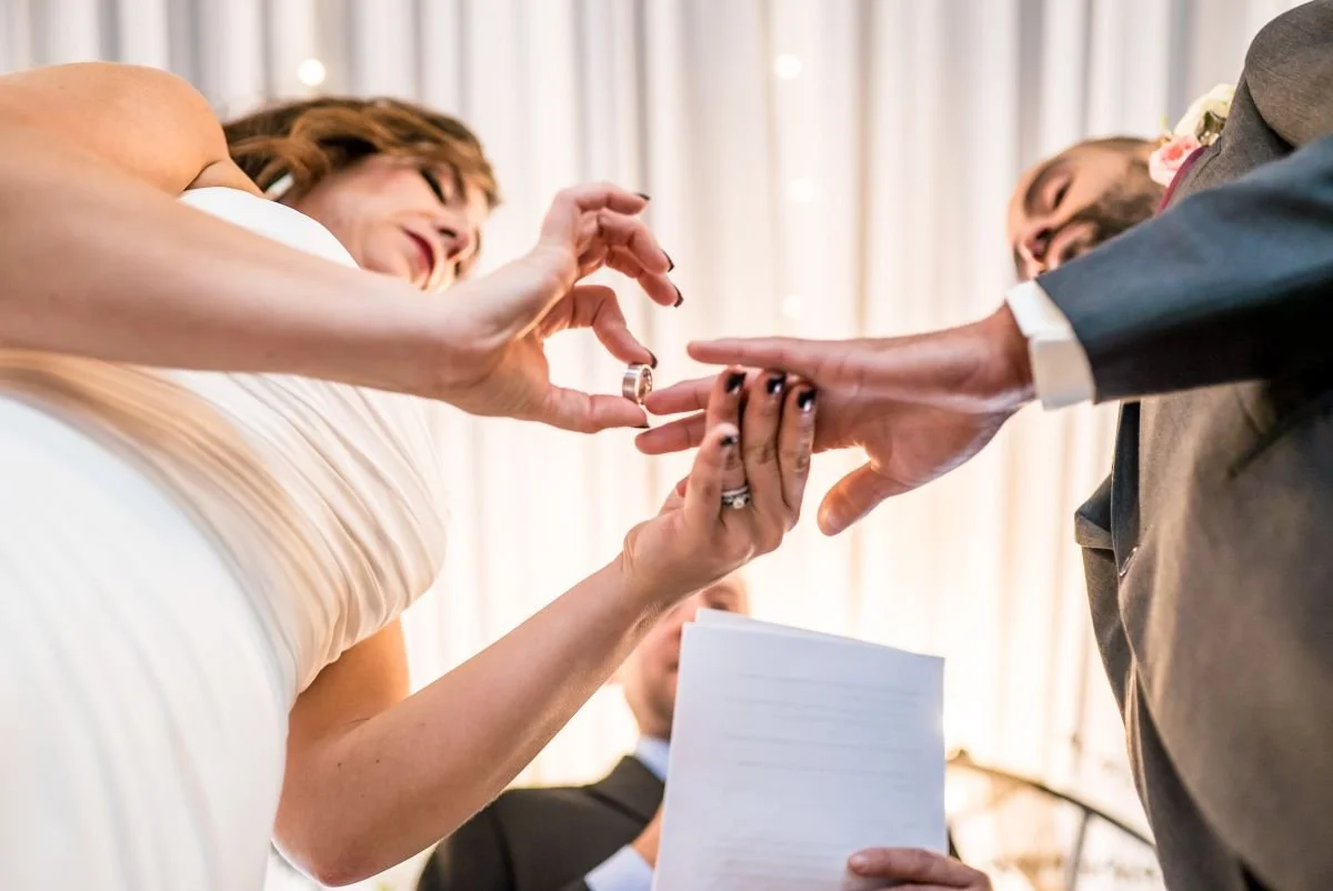 A bride in a white dress looks down at the groom's hand while she places his wedding ring on his finger at Ashgrove Acres in Manitoba.