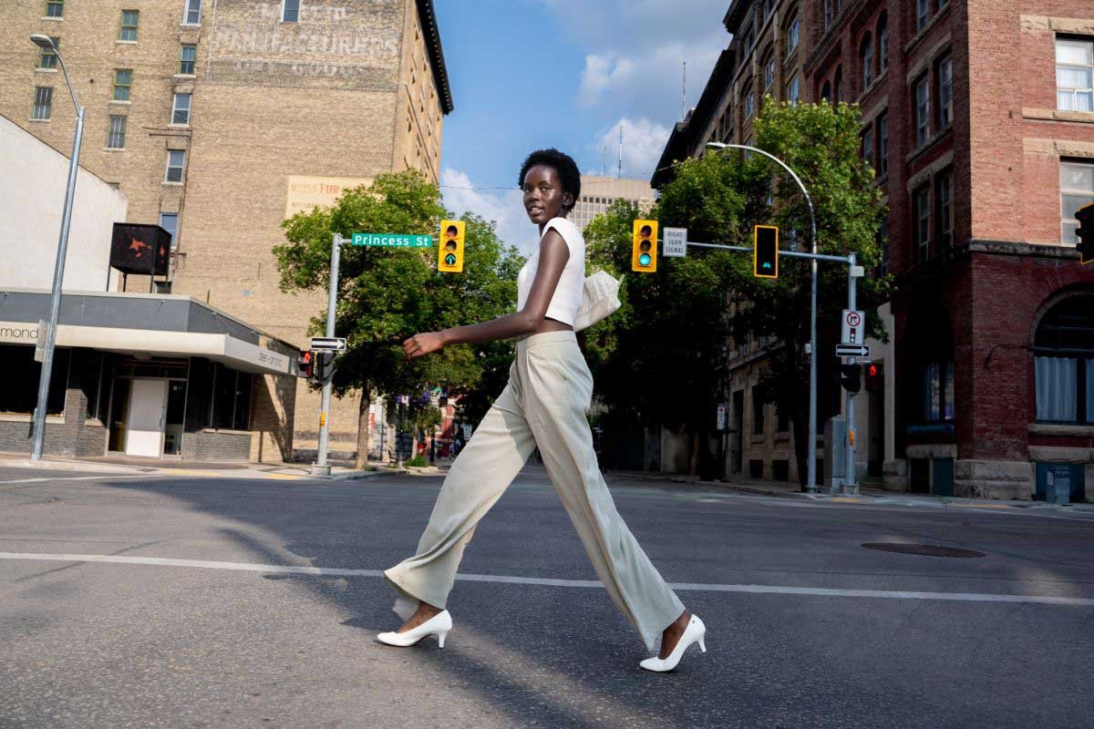A female fashion model in a white sleeveless crop top and beige trousers walks across a city crosswalk at Princess Street in Winnipeg Manitoba.