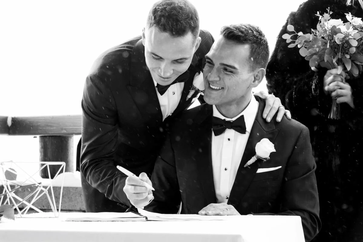 Candid black and white moment of two grooms in elegant black tuxedos smiling as they sign wedding documents at a snowy mountain venue.