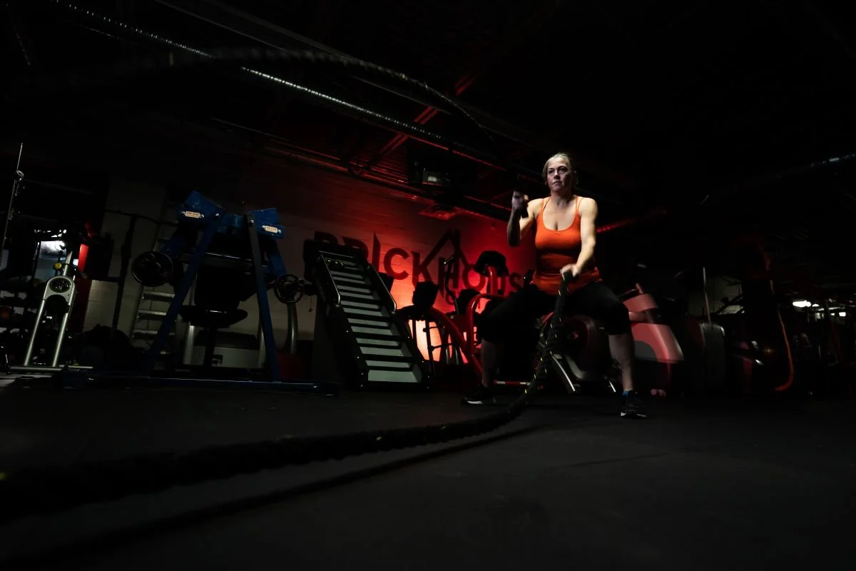 A dynamic, low-key commercial print advertisement photograph of a woman in an orange tank top slamming battle ropes inside Brick House Gym in Winnipeg, captured for a Fitness Manitoba promotional campaign.