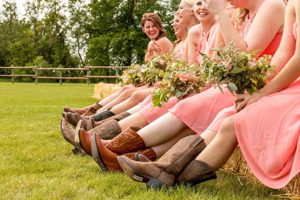 Bridesmaids in coral pink dresses wearing leather cowboy boots sitting on hay bales at a rustic Hitch 'N Post country wedding in Manitoba.