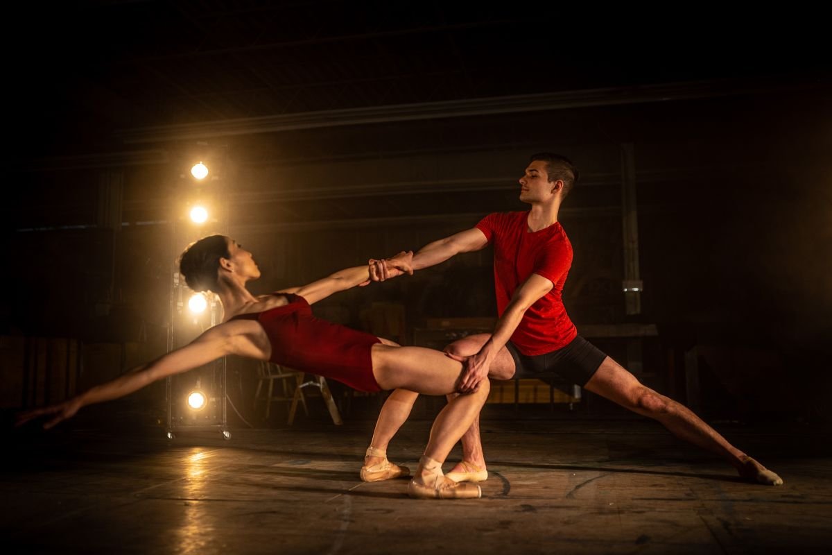 A dramatic creative editorial photograph of two ballet dancers performing a dynamic duet pose on a stage illuminated by a warm vertical light stand, shot for the Royal Winnipeg Ballet by a Winnipeg dance photographer.
