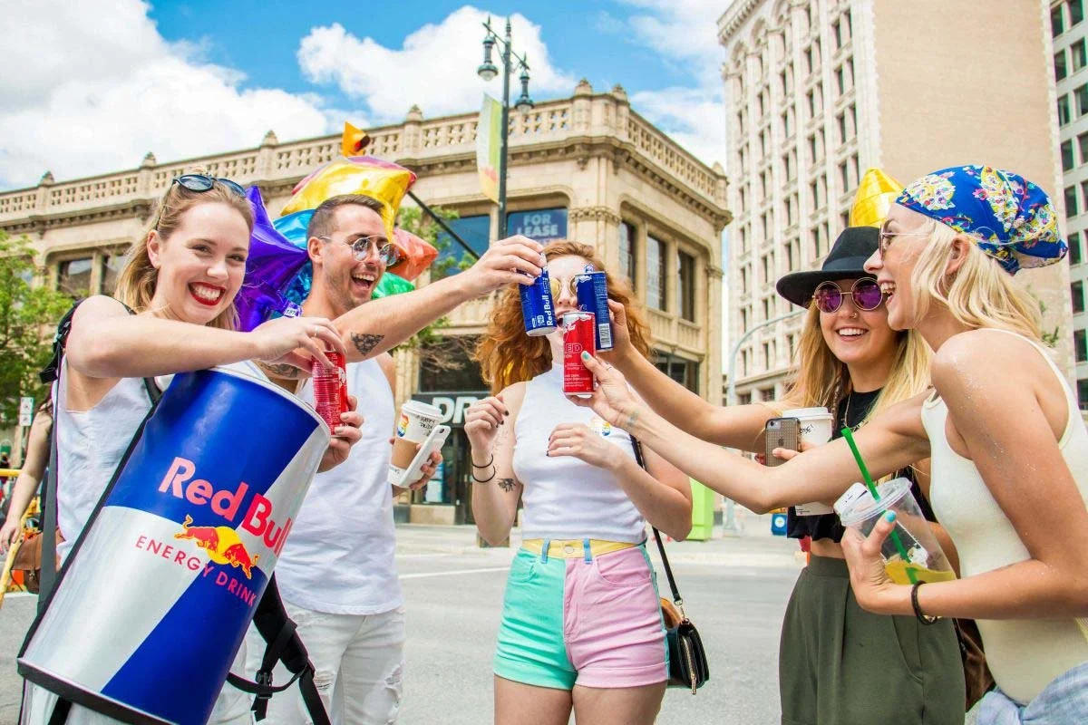 A vibrant commercial event photograph of people cheering with Red Bull energy drink cans during the Pride Winnipeg festival, captured on Portage Avenue by a Winnipeg commercial photographer.