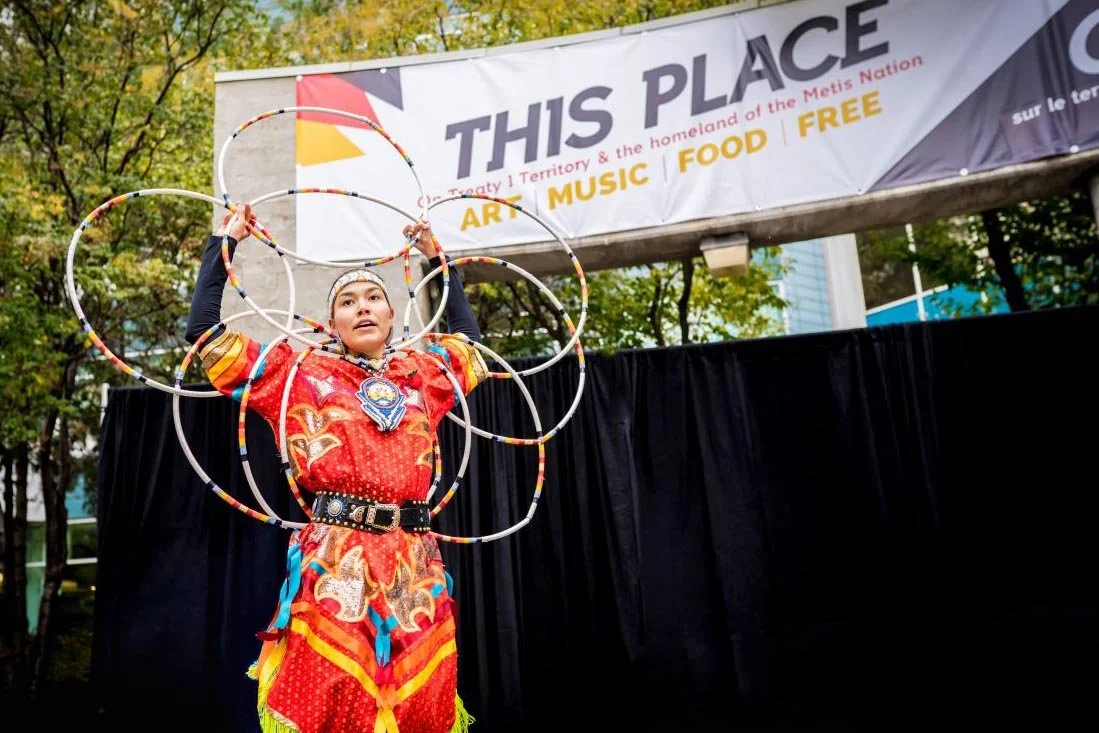 An Indigenous hoop dancer in vibrant traditional regalia performs on stage with multiple hoops at a Winnipeg Arts Council public art grand opening event, underneath a banner reading This Place: On Treaty 1 Territory and the homeland of the Metis Nati