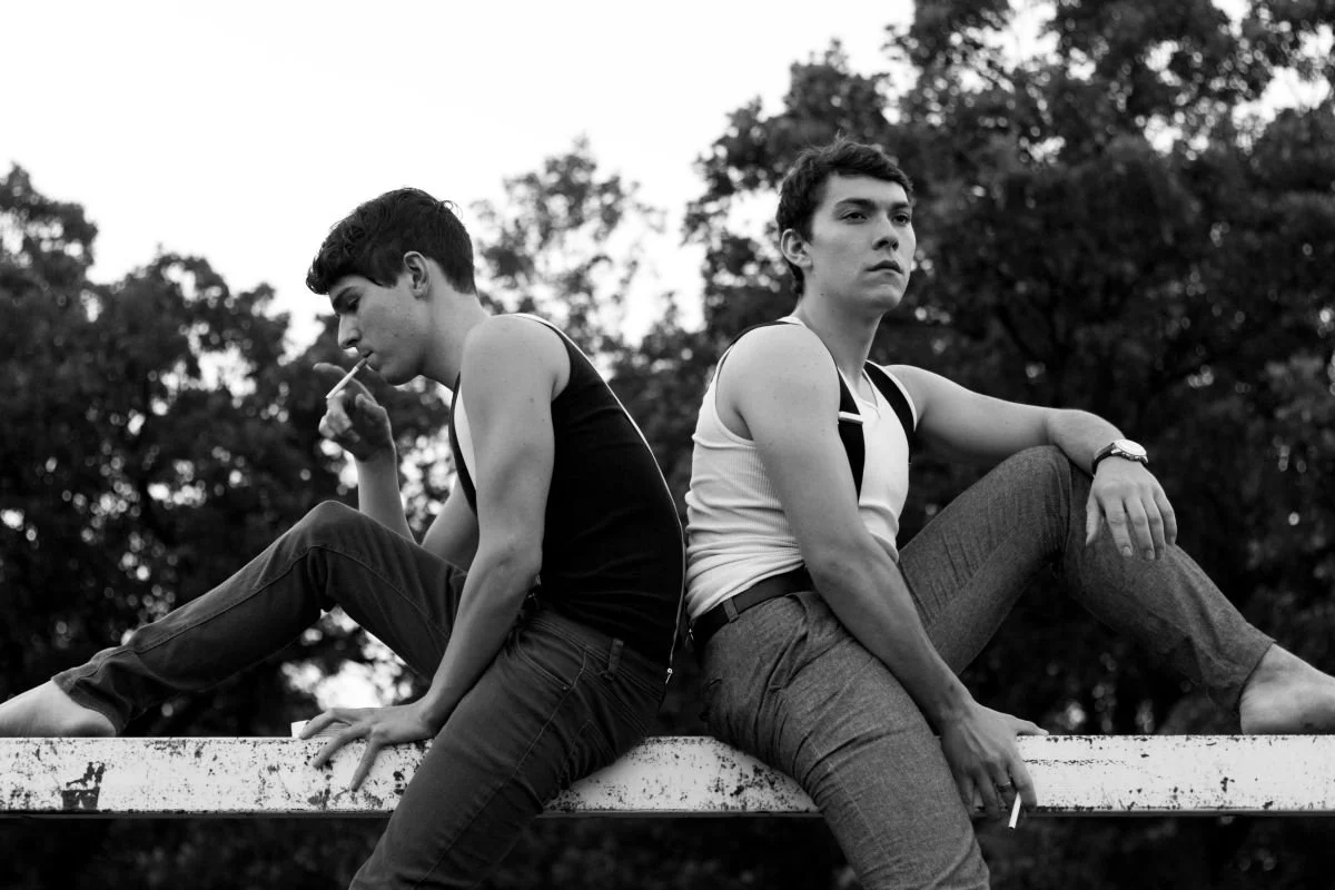 A dramatic black and white men's fashion editorial photo of two young men in tank tops sitting back-to-back on a metal railing at Assiniboine Park in Winnipeg.