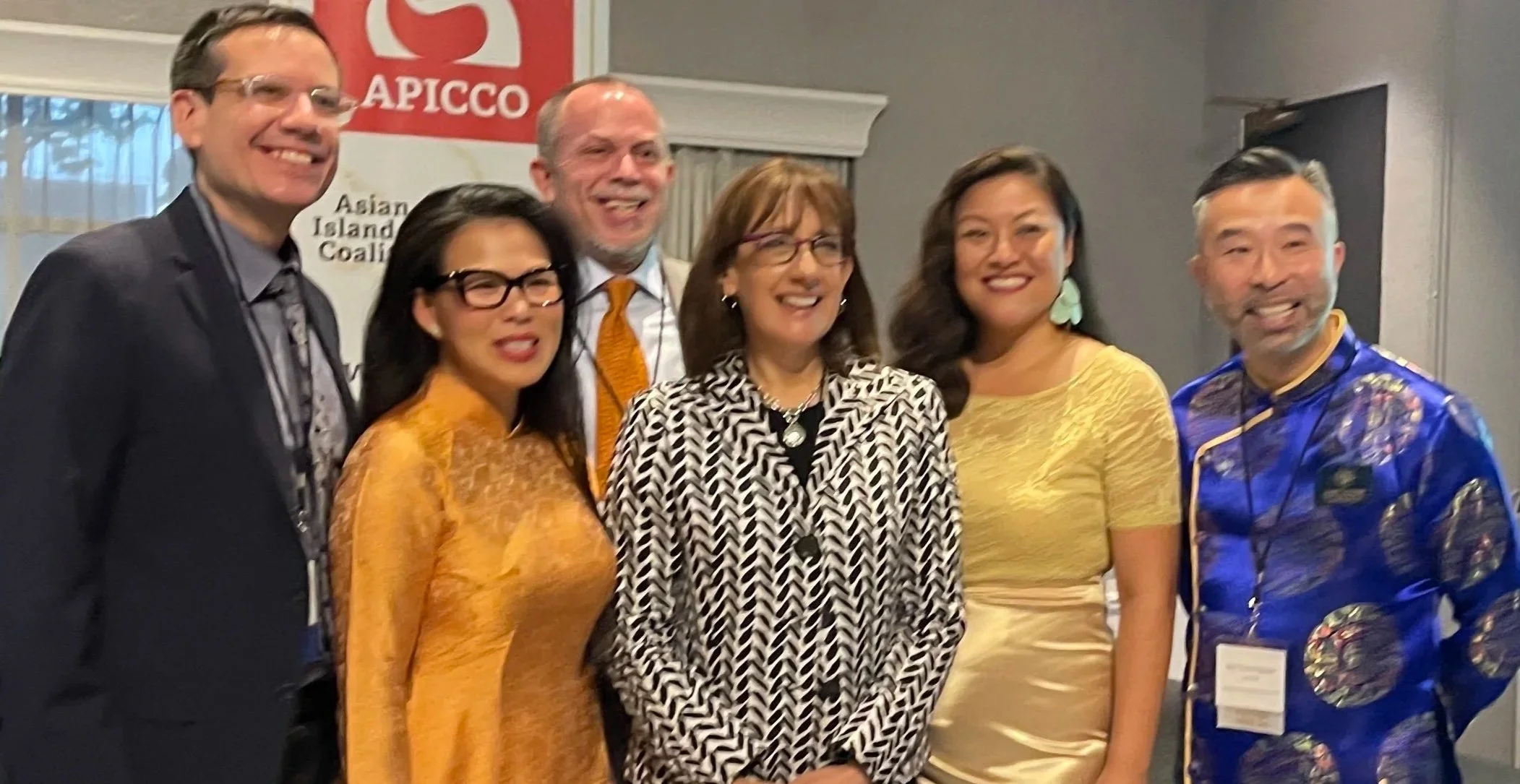 Group of six diverse people smiling and posing for a photo at an event, with a banner in the background that says 'APICCO' and 'Asian Island Coalition'.