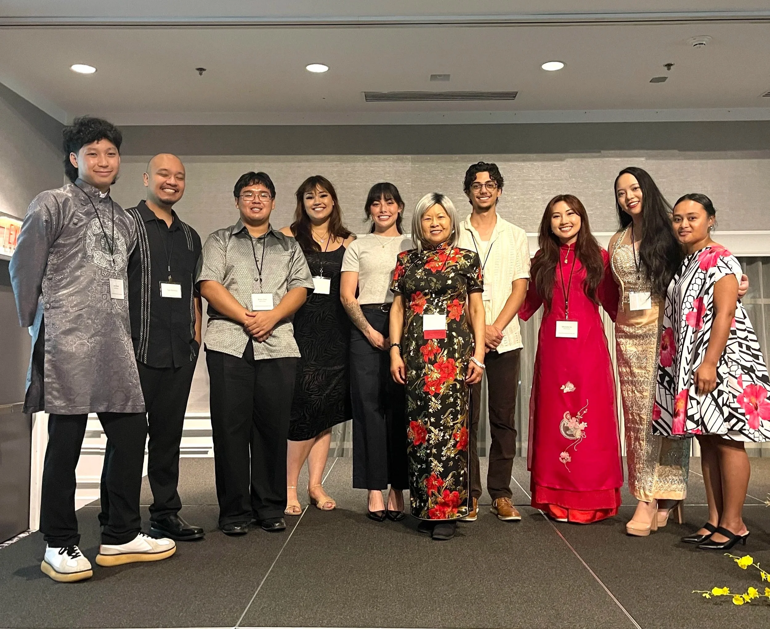 Group of diverse people standing together indoors, smiling for a photo, with some wearing traditional and modern attire, and name tags around their necks.