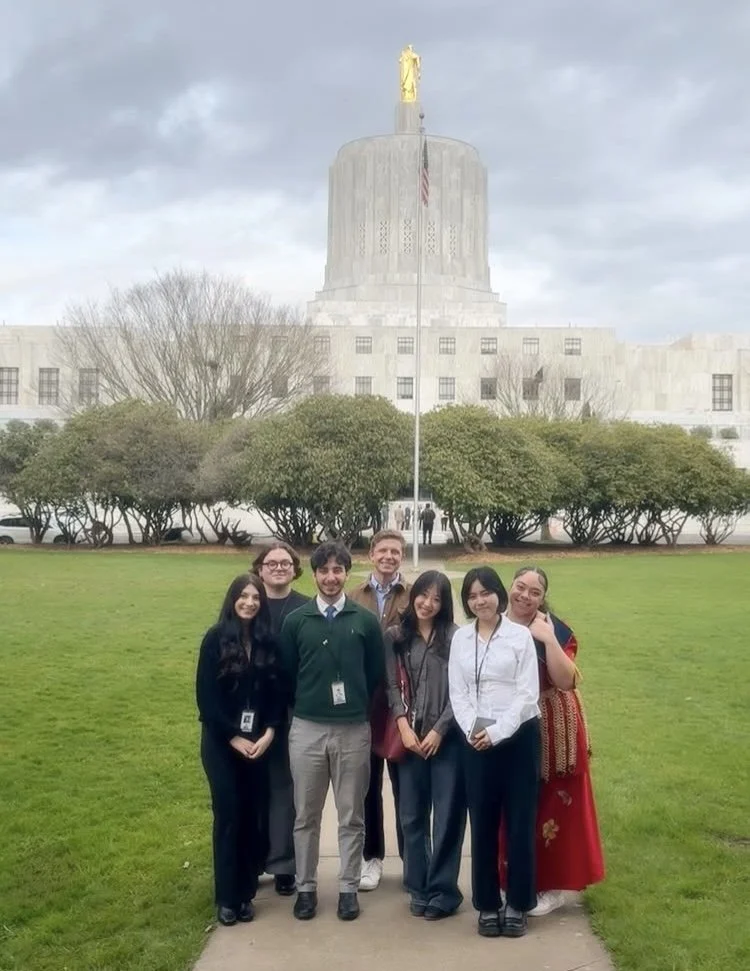 Group of seven diverse people standing on a sidewalk in front of a grassy area with trees and a large white government building with a dome and a statue on top, under a cloudy sky.
