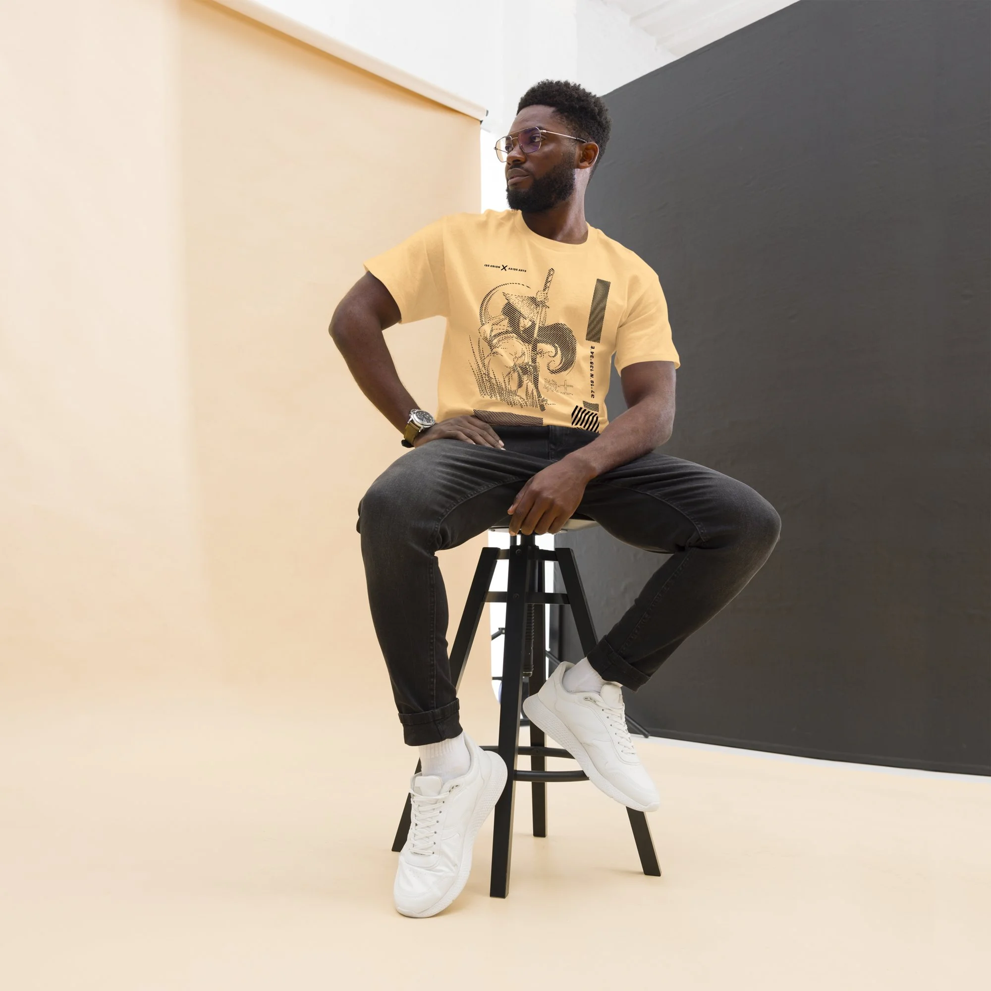 A man with glasses and a beard sitting on a tall black stool against a dual-colored background (white and dark gray) in a studio setting, wearing a yellow graphic T-shirt, dark jeans, white sneakers, and a wristwatch, looking to his left.
