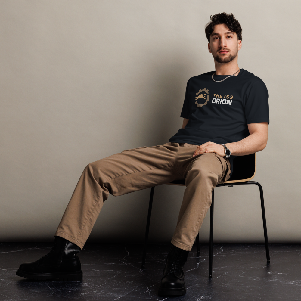 A young man with dark wavy hair, beard, and mustache, sitting on a black chair against a plain light-colored wall. He is wearing a black t-shirt with a logo and text, beige pants, black combat-style boots, a watch, a necklace, and a bracelet, looking directly at the camera.