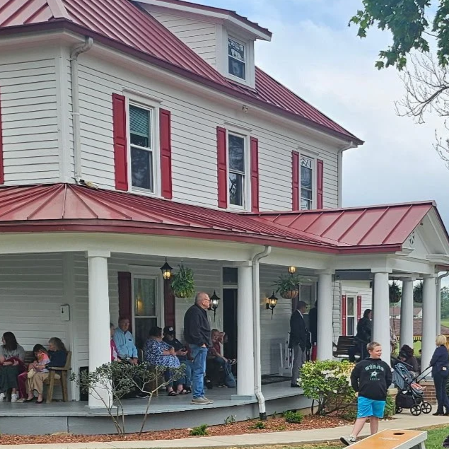 People gathered on the porch of a white house with a red metal roof, featuring red window shutters, during daytime.