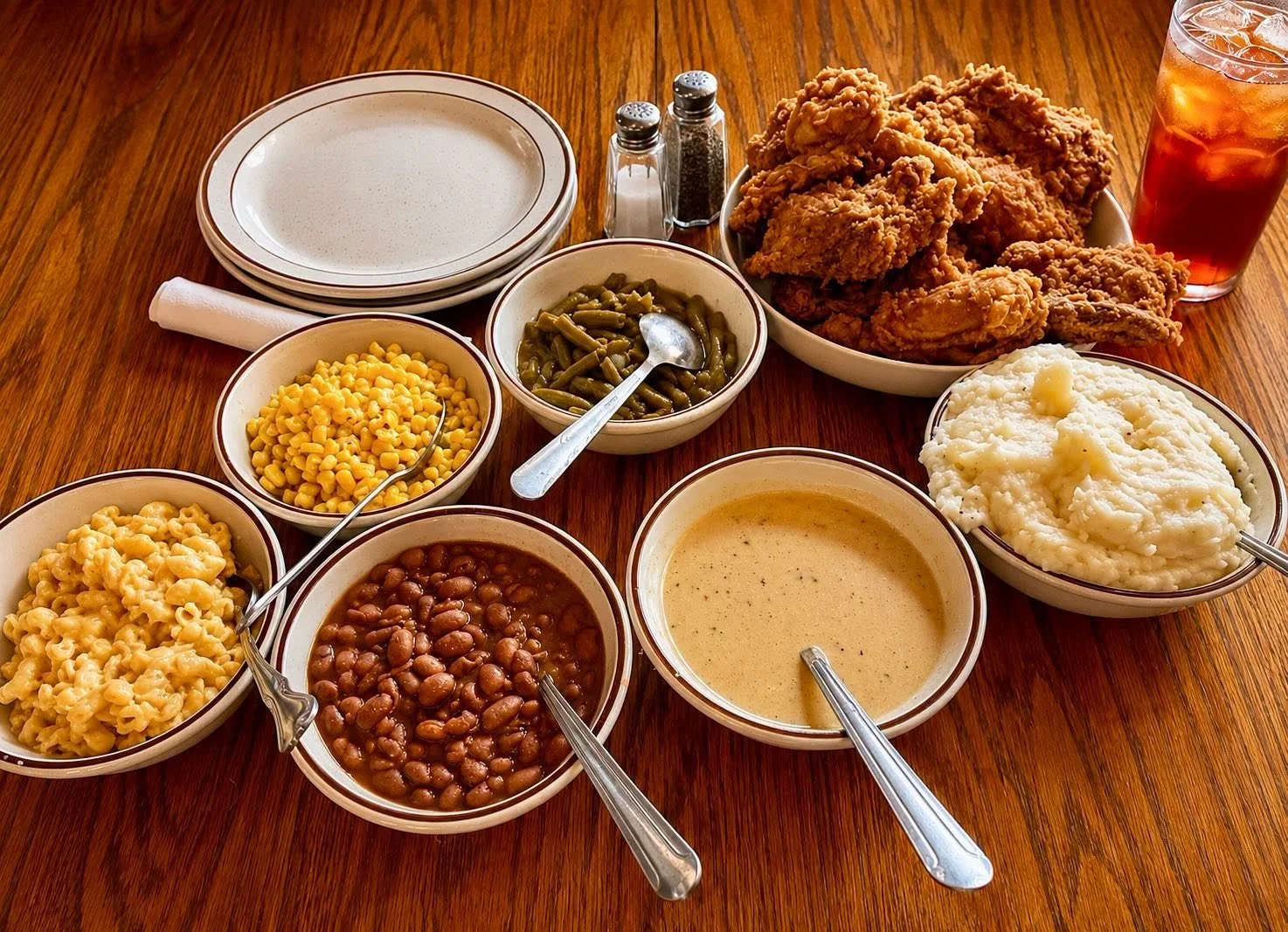 A table with various bowls of Southern comfort foods, including mashed potatoes, fried chicken, green beans, corn, baked beans, macaroni and cheese, and iced tea.
