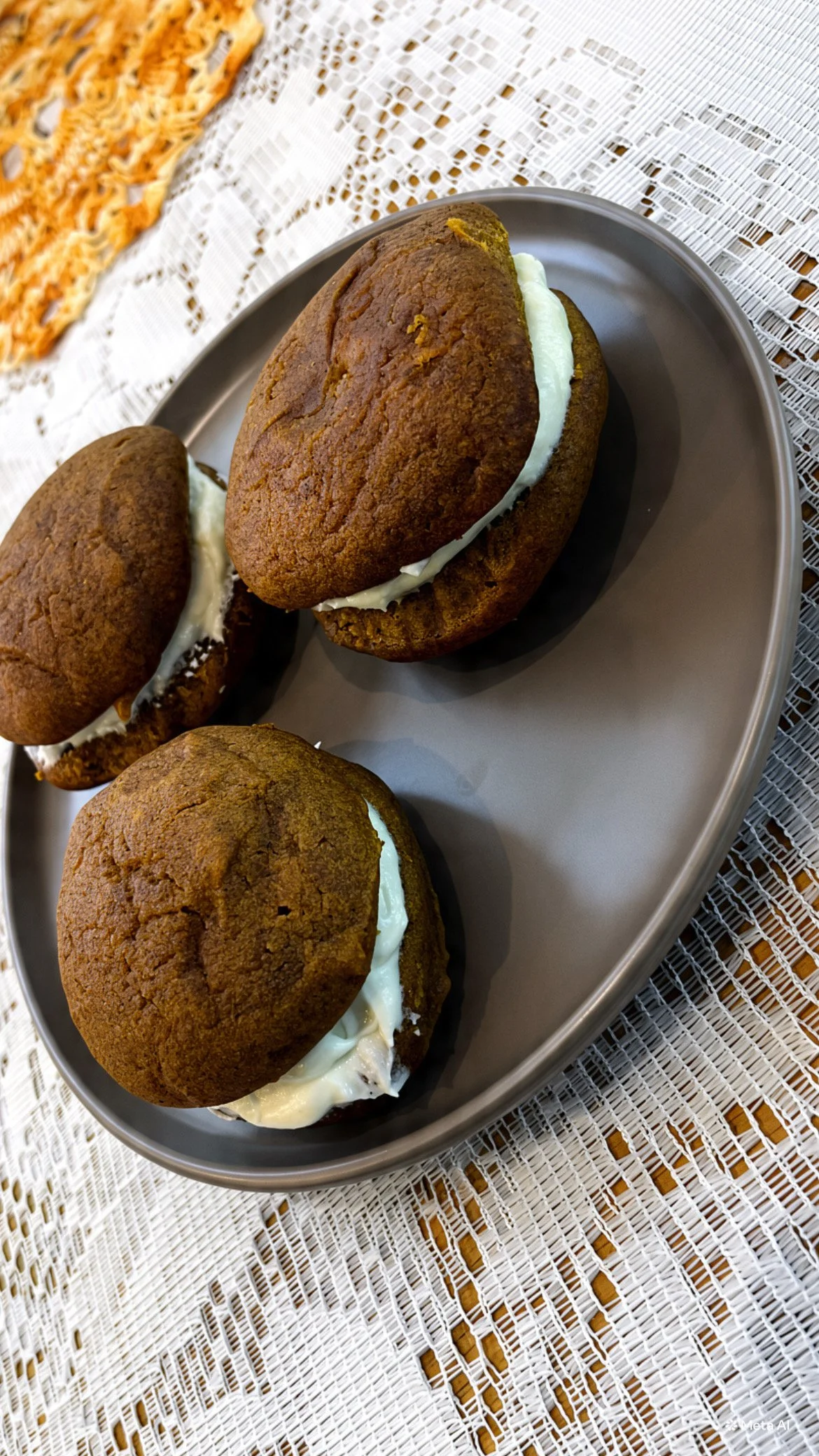 Three pumpkin whoopie pies with cream cheese filling on a gray plate, placed on a lace tablecloth.