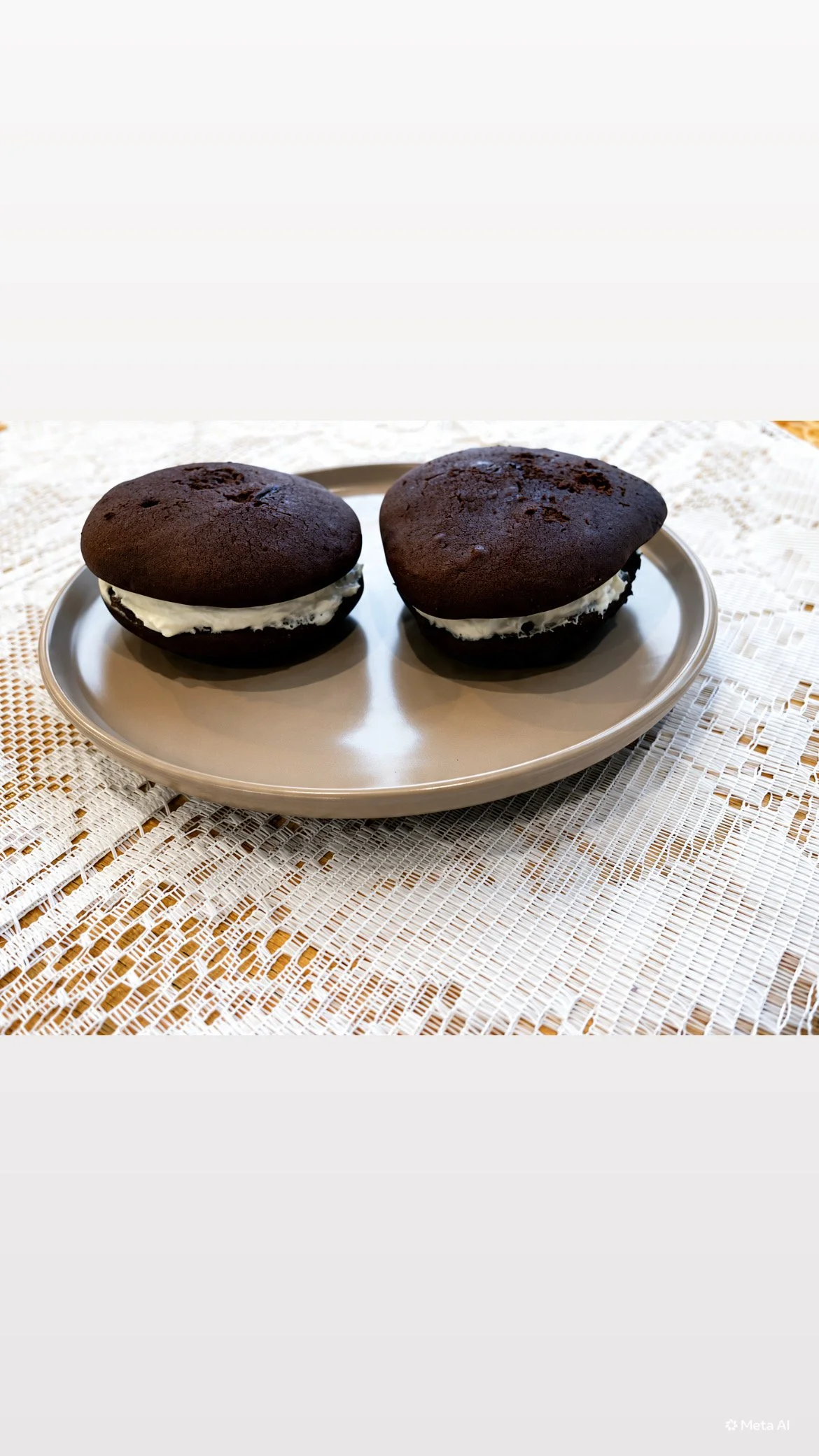 Two black whoopie pies with white cream filling on a silver plate, placed on a white woven lace cloth.
