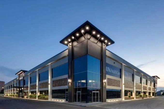 Modern two-story medical office building with blue and gray exterior, large glass windows, and a sign reading 'UK Healthcare' at the corner, taken at dusk.