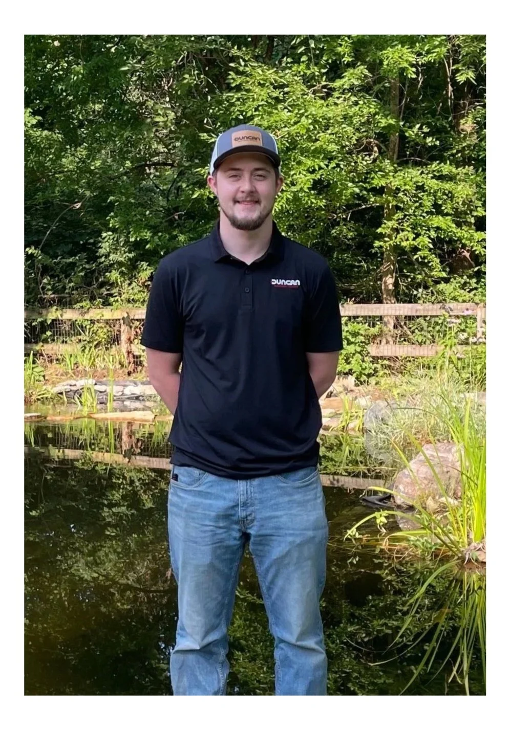 A young man standing outdoors near a pond with green foliage and a wooden fence in the background. He is wearing a black shirt with a logo, blue jeans, and a cap.