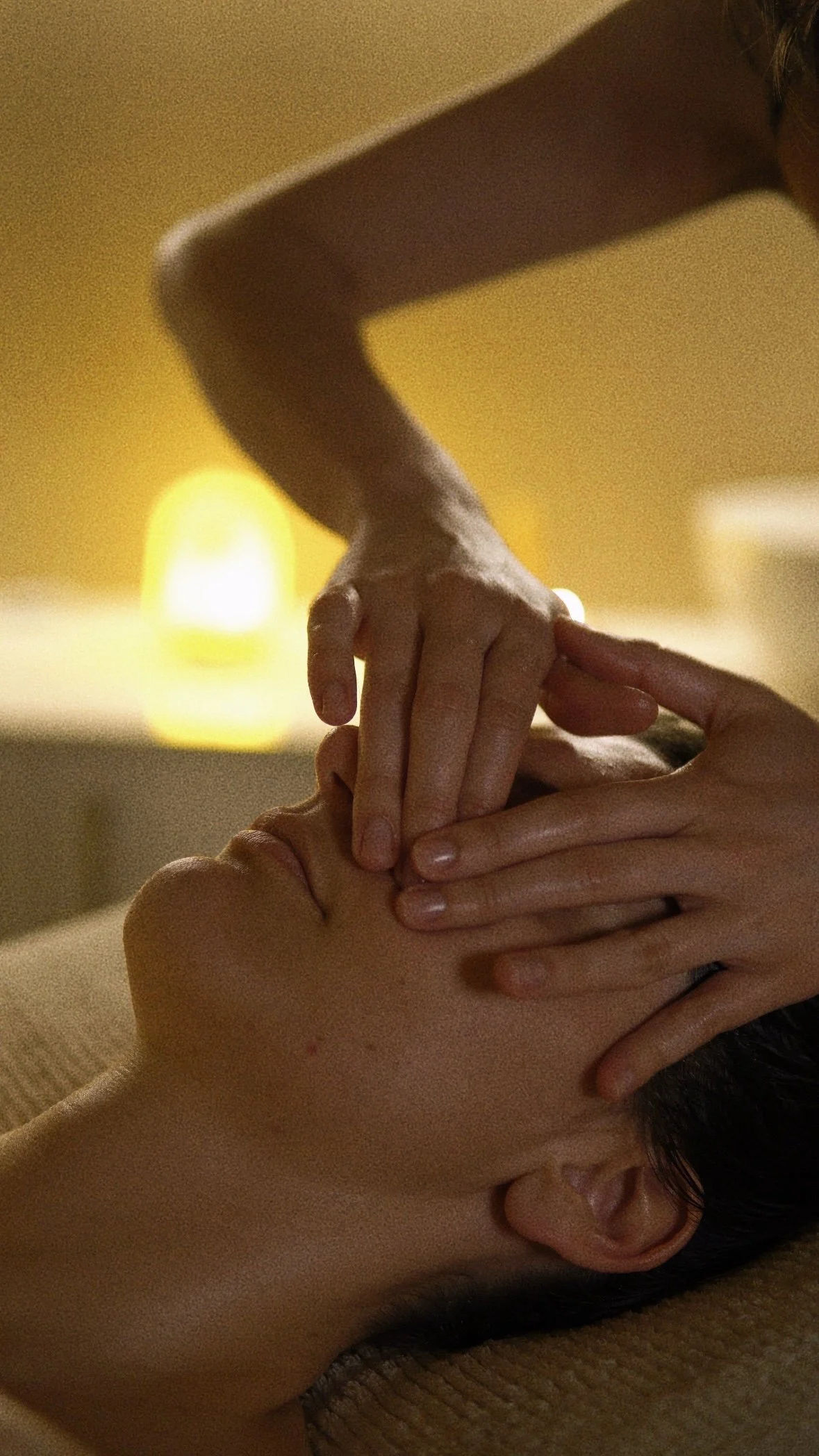 Person receiving a massage with hands applying pressure on their face and neck, in a dimly lit room.