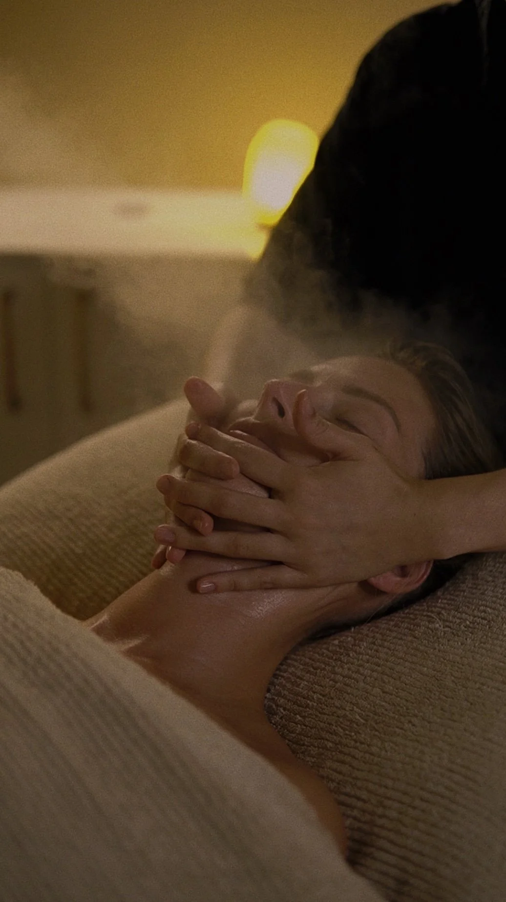 A person receiving a facial massage while lying down, with a facial steamer over the top for a luxury experience. The background is softly lit, creating a warm and calming atmosphere.