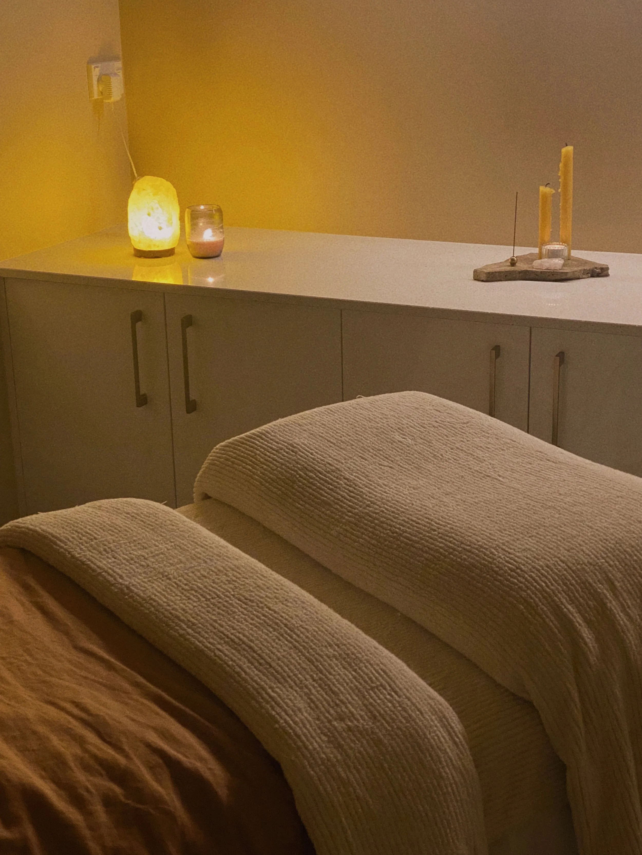 A modern, cozy facial room with a comfortable contoured bed, warm blanket. The background has dimmed light, softly lit by a salt lamp creating a warm and calming atmosphere.