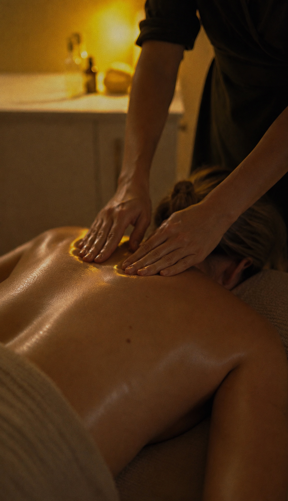 A person receiving a back massage in a dimly lit room, lying face down on a massage table while a massage therapist applies oil on their back.