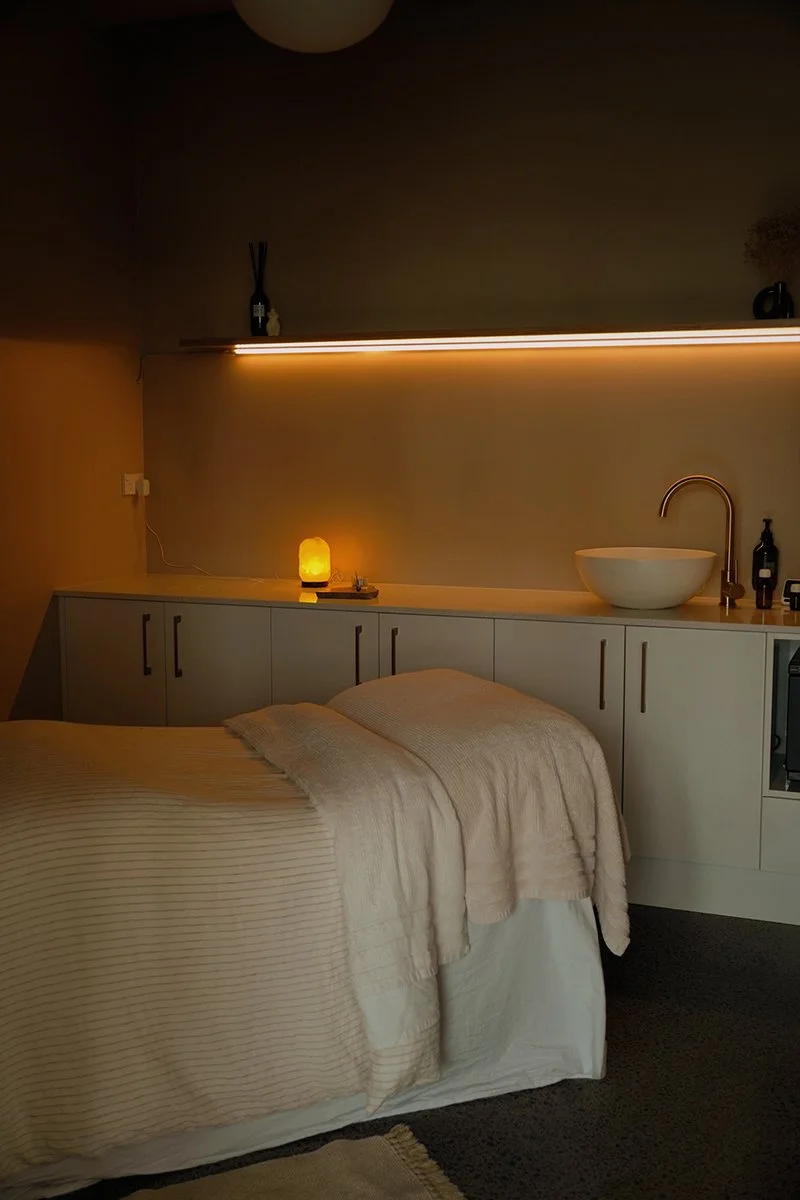 A modern, cozy facial room with a comfortable contoured bed, warm blanket. The background has dimmed light, softly lit by a salt lamp creating a warm and calming atmosphere.