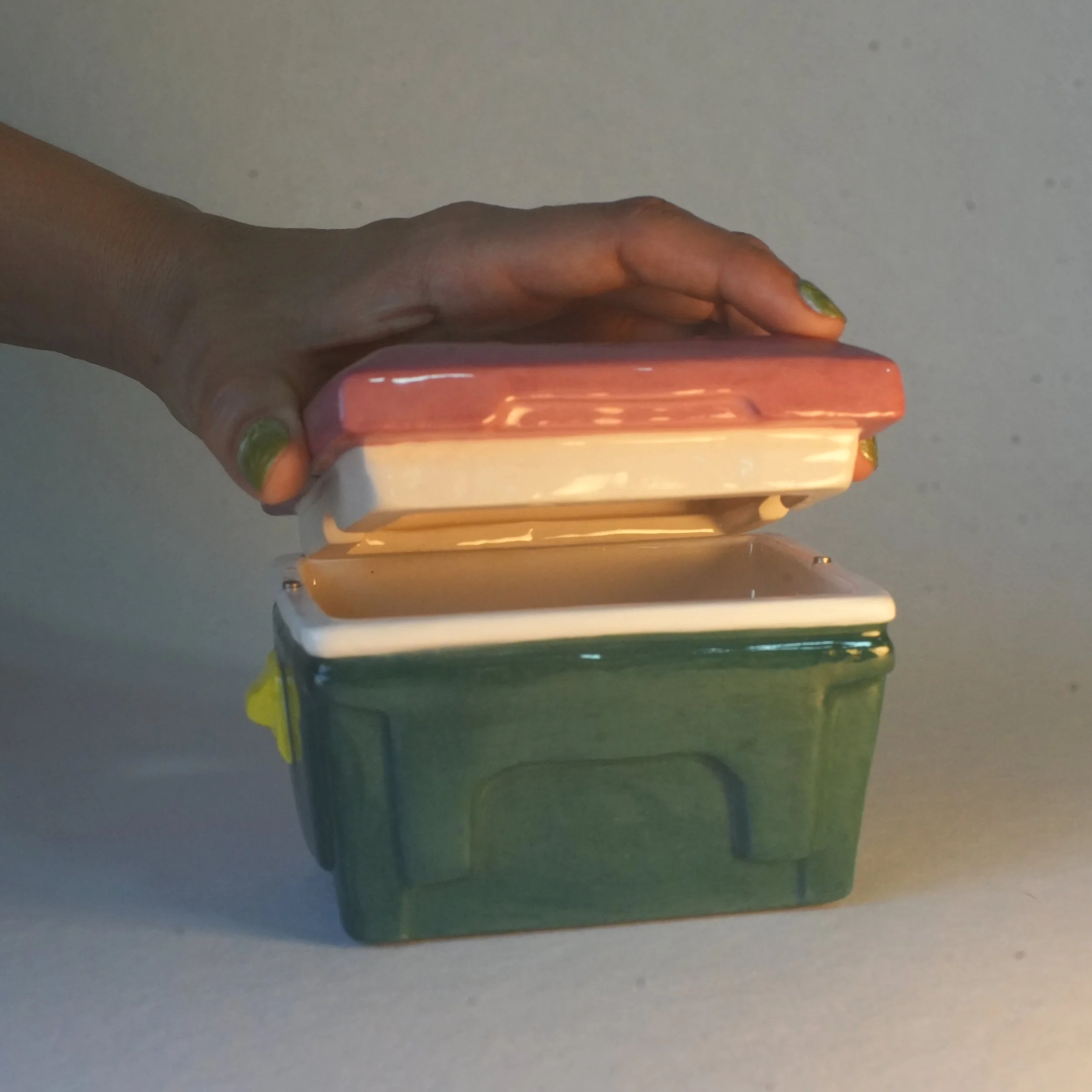Close-up of a person's hand with painted nails holding a retro cooler inspired trinket box and ashtray lid against a neutral background.