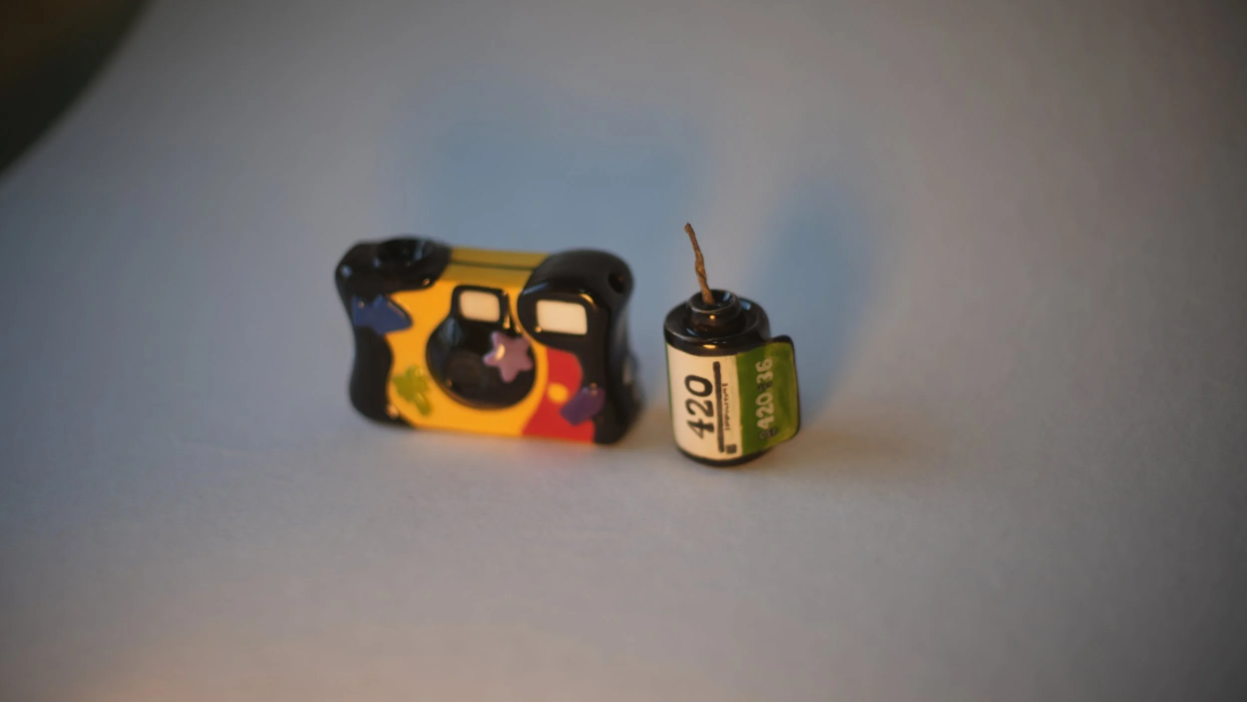 A ceramic disposable camera inspired ceramic pipe with various sticker-like designs, next to a 35mm camera roll inspired hemp wick holder in green and white with shell with the number 420 labeled on it.