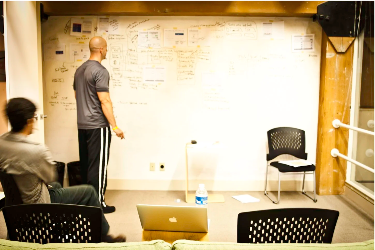 A man writing on a whiteboard with notes and diagrams while two people sit and listen, in an office or conference room.
