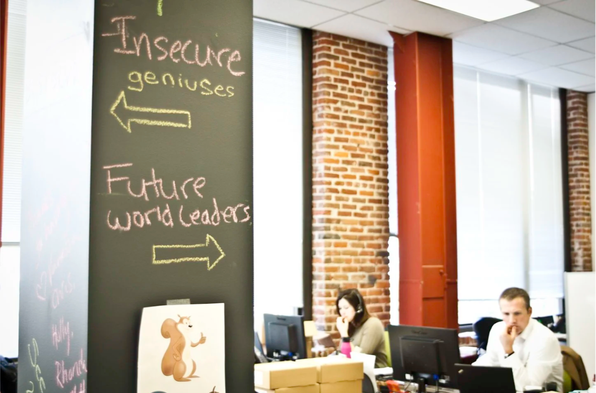 Office space with a chalkboard wall that has handwritten messages in different colors and arrow signs. There are two people working at desks in front of a brick wall with large windows.