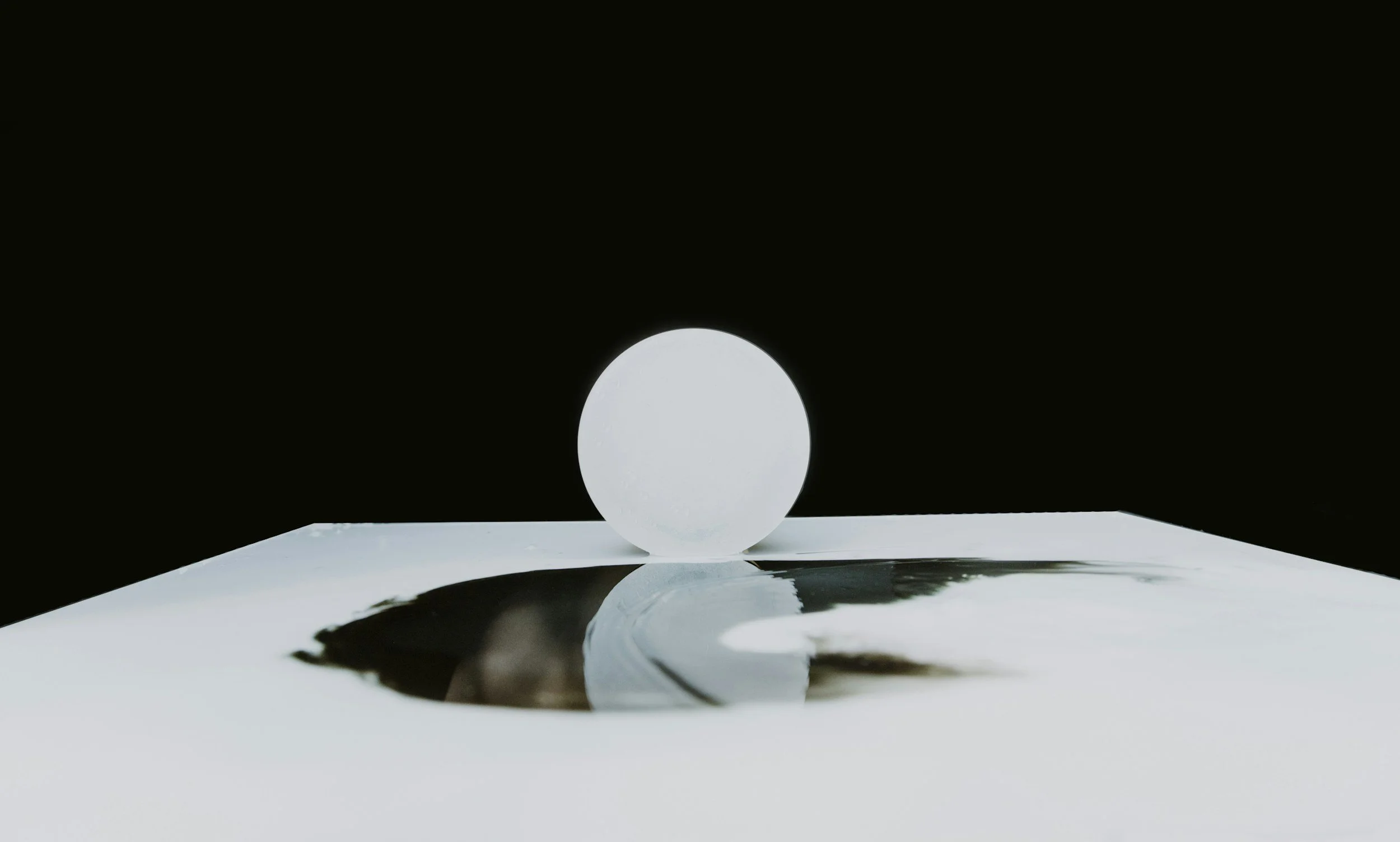 A white sphere on a reflective surface with black background, casting a shadow.
