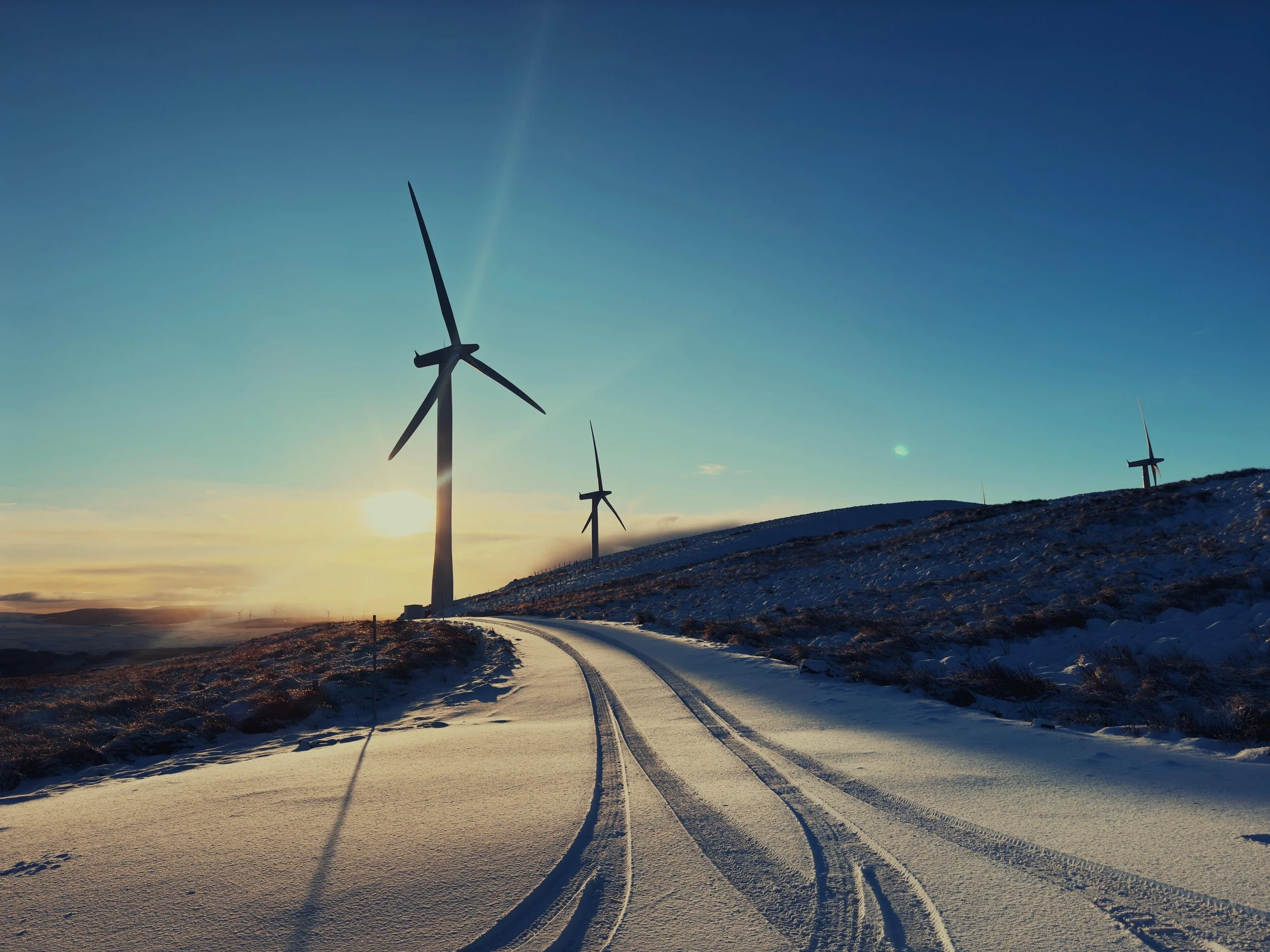 A scenic winter landscape featuring a snow-covered road leading towards wind turbines on a hill at sunset or sunrise, with a clear blue sky and faint clouds.