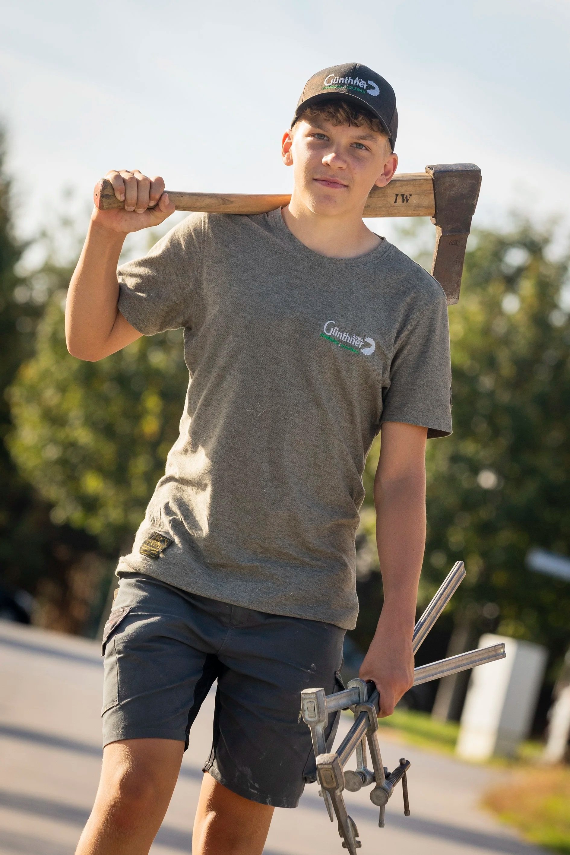 Junger Mann trägt schwarze T-shirt und kurze Hosen, hält eine Elektrosäge in der Hand, trägt eine schwarze Kappe mit Logo, trägt eine Smartwatch, steht im Freien bei gutem Wetter.