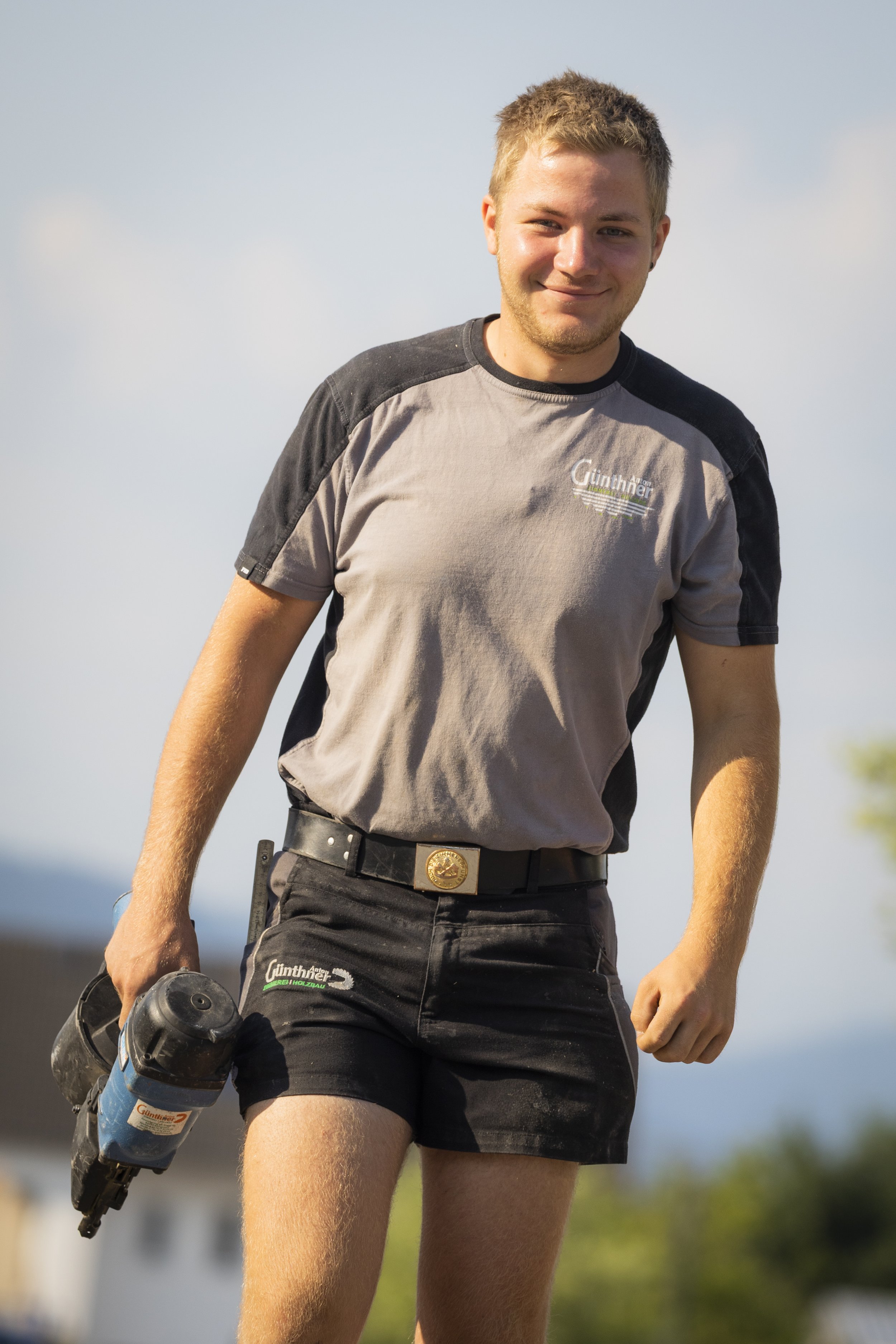 Ein junger Mann mit kurzen Haaren und einem leichten Bart, trägt ein graues T-Shirt mit schwarzen Schultern, schwarze Shorts, einen Gürtel mit einer goldenen Schnalle und hält einen Bohrhammer in der rechten Hand. Er steht im Freien bei sonnigem Wetter, im Hintergrund sind Blüten und Bäume sichtbar.
