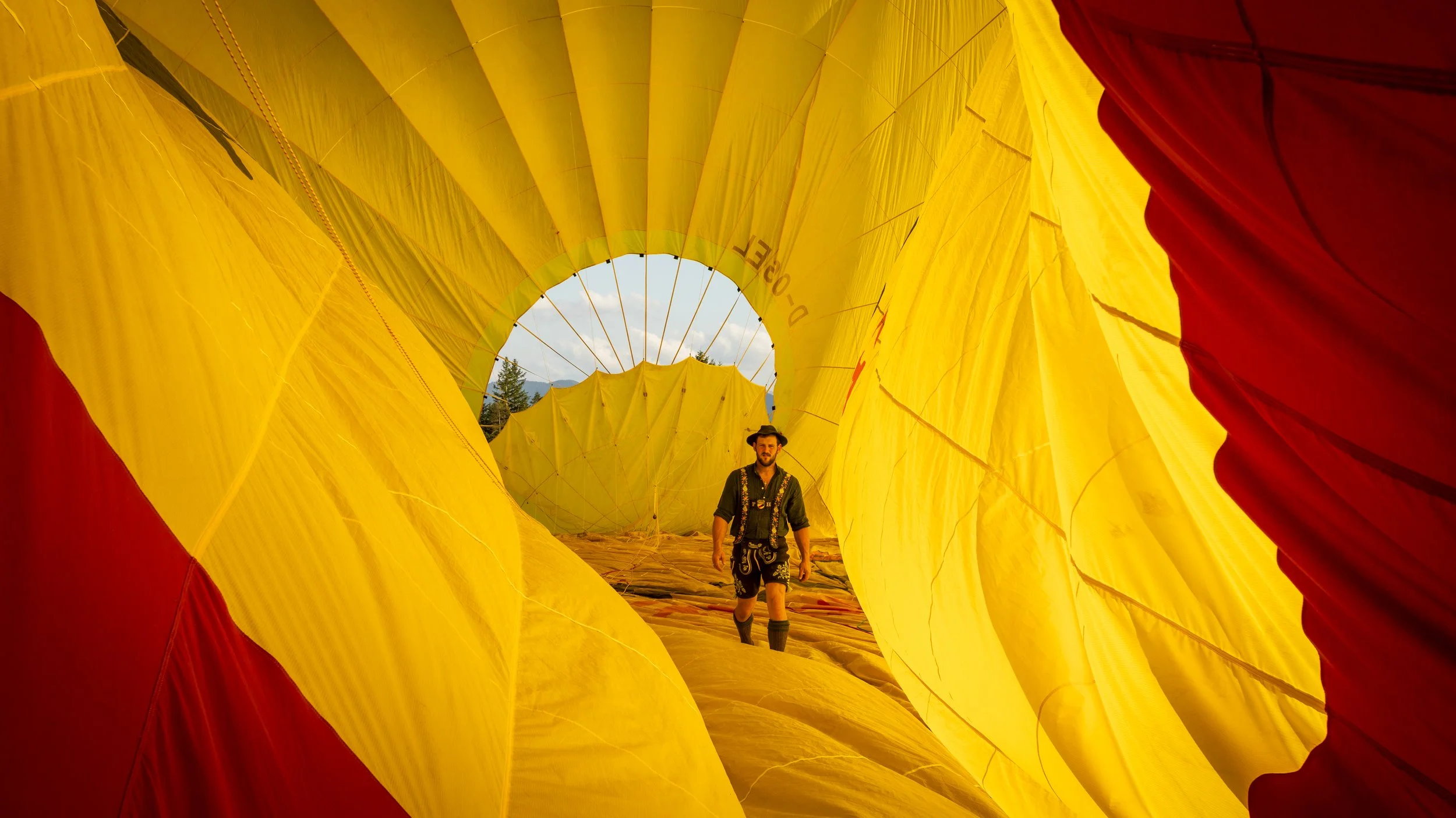 Blick in einen aufblasbaren Heißluftballon, mit einem Mann in traditionellen Kleidern und Hut im Inneren, während die Hülle aufgeblasen wird.